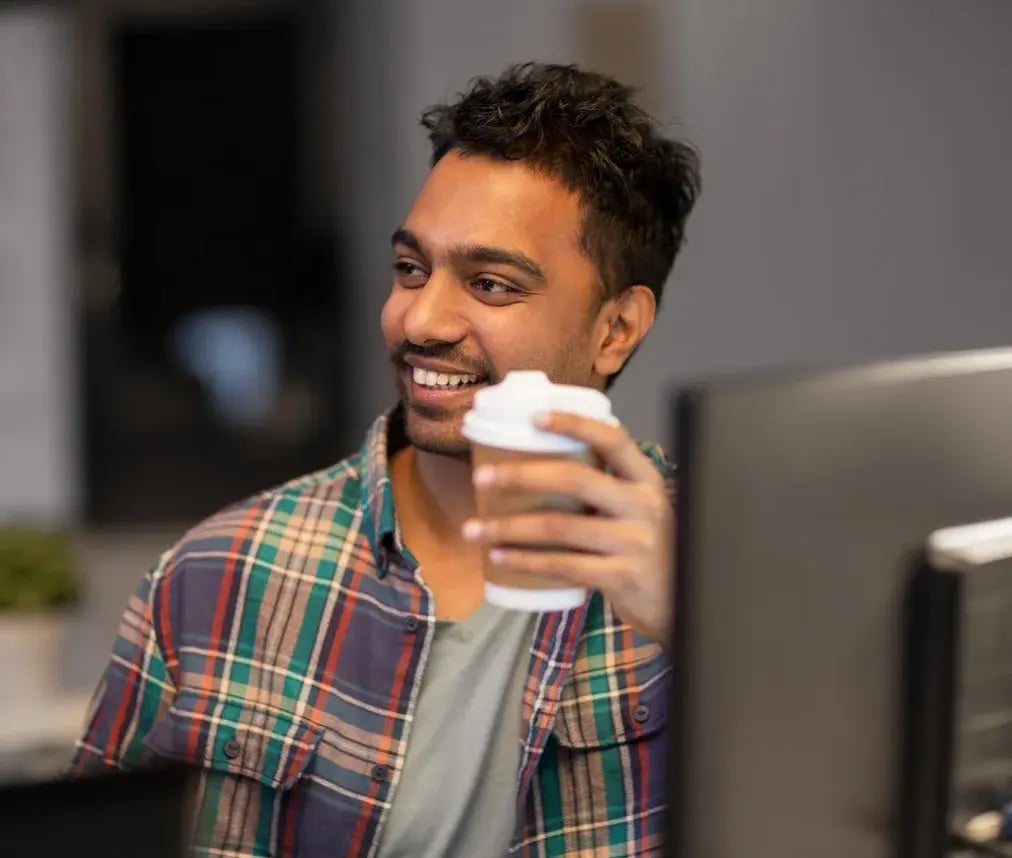 A smiling office worker holding a takeaway coffee beside his computer monitor