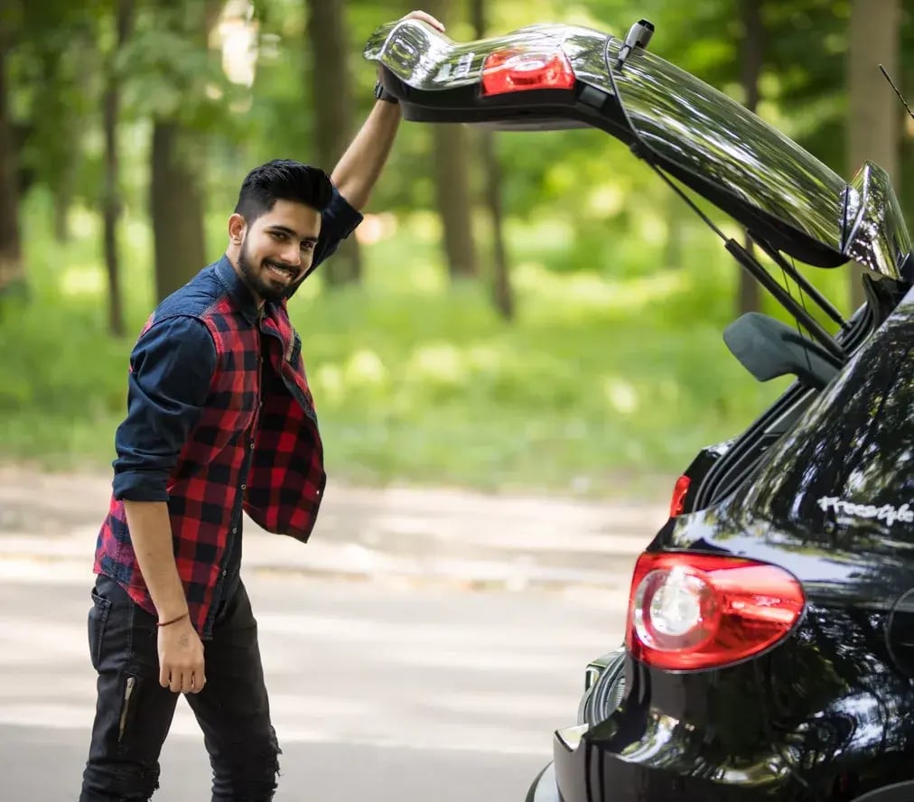 A smiling man lifting the boot lid of his car on a tree-lined road