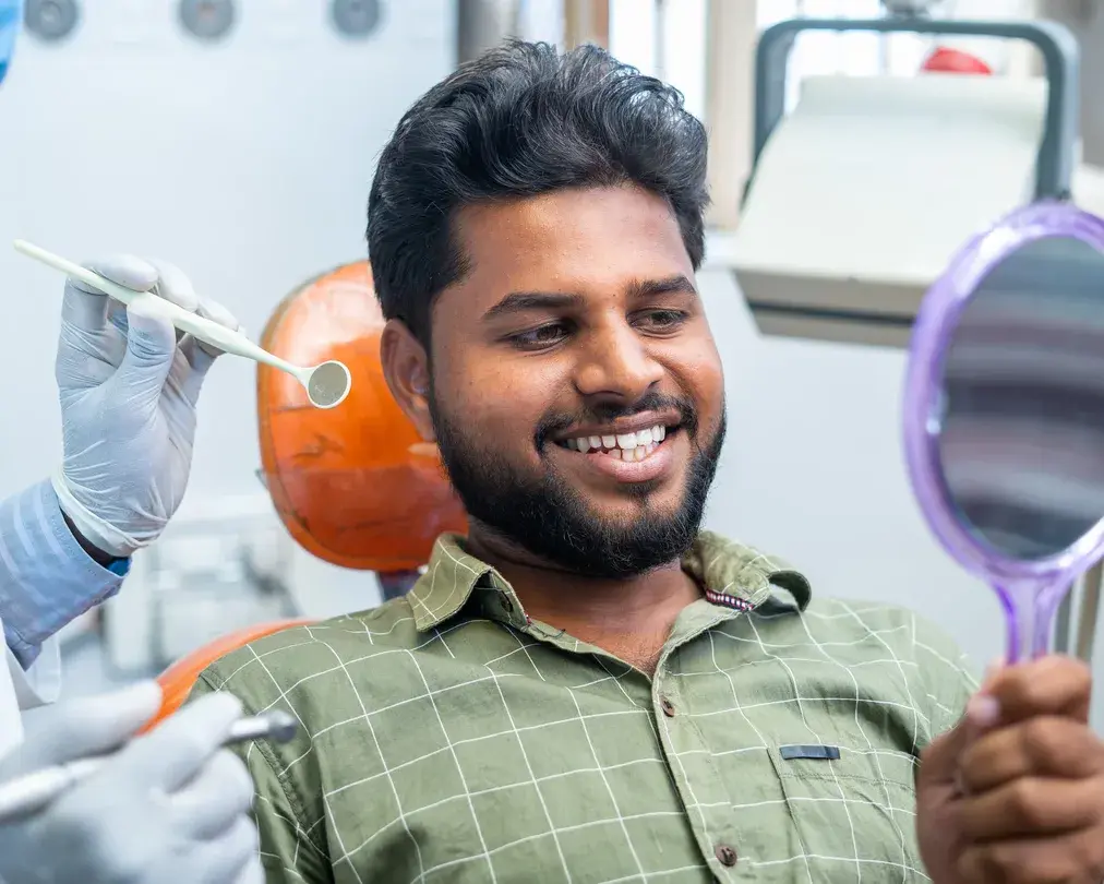 A smiling patient checking his teeth in a hand mirror while the dentist holds a dental mirror beside him