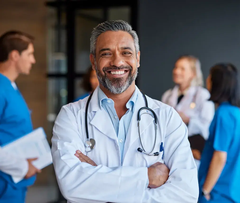A smiling doctor in a white coat with a stethoscope, colleagues talking in the background