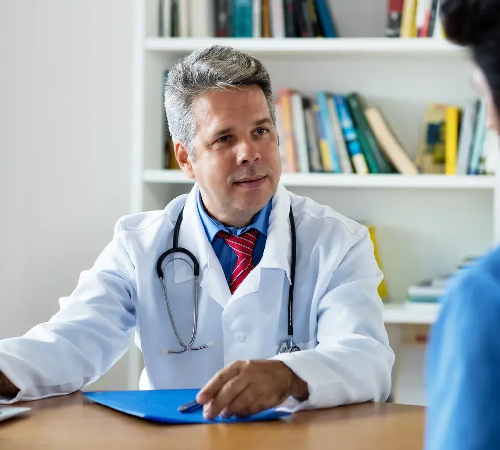A doctor in a white coat talking with a patient across a desk at a German practice